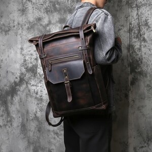 May include: A dark brown leather backpack with a roll-top closure and multiple pockets. The backpack has adjustable straps and a front flap with a buckle. The leather has a distressed finish, giving it a vintage look. The backpack is being worn.