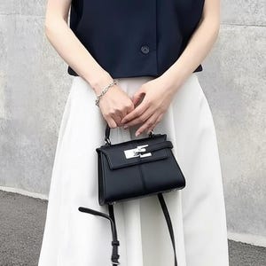 May include: A black handbag with a silver clasp is held. The bag has a structured design and a long, thin strap. The person is wearing a dark blue top and a white skirt. A silver bracelet is on the person's wrist. The background is a grey wall.