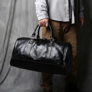 Puede incluir: Una bolsa de viaje de cuero negro con un acabado brillante. La bolsa tiene dos asas superiores y una correa para el hombro. La bolsa es sostenida por una persona. El fondo es gris neutro.