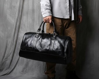 Puede incluir: Una bolsa de viaje de cuero negro con un acabado brillante. La bolsa tiene dos asas superiores y una correa para el hombro. La bolsa es sostenida por una persona. El fondo es gris neutro.