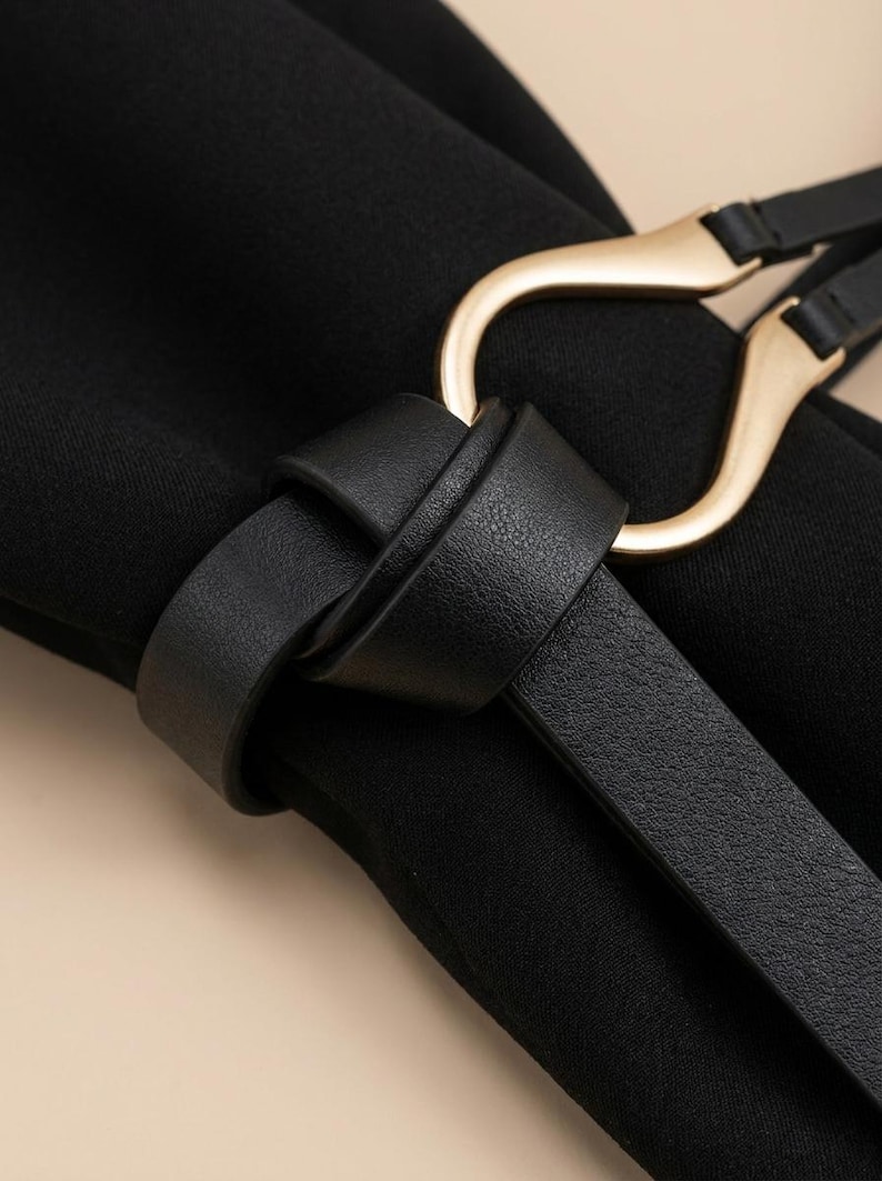 Puede incluir: Primer plano de un cintur&oacute;n negro con hebilla dorada. El cintur&oacute;n est&aacute; hecho de un material liso similar al cuero y est&aacute; anudado. La hebilla tiene un dise&ntilde;o curvo &uacute;nico.