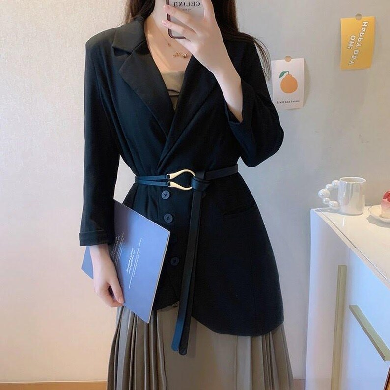 Puede incluir: Un blazer negro con cintur&oacute;n de hebilla dorada, combinado con una falda plisada. El blazer tiene un dise&ntilde;o asim&eacute;trico &uacute;nico y est&aacute; abotonado. Un libro azul se sostiene en la mano izquierda.