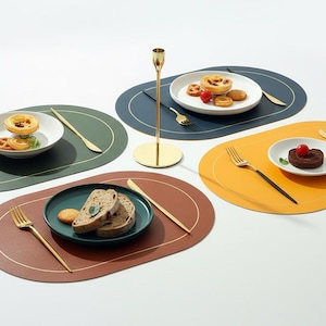Puede incluir: Vista aérea de cuatro manteles individuales ovalados en verde, azul marino, marrón y amarillo, cada uno con un borde dorado. Cada mantel individual tiene un plato, cubiertos y comida. Un candelabro dorado está en el centro.