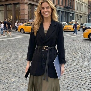 Puede incluir: Una persona con una blazer negra con cintur&oacute;n a juego y una falda plisada larga verde oliva. La blazer tiene cierre de botones y se combina con un top oscuro. El fondo muestra una calle de la ciudad con taxis amarillos y peatones.
