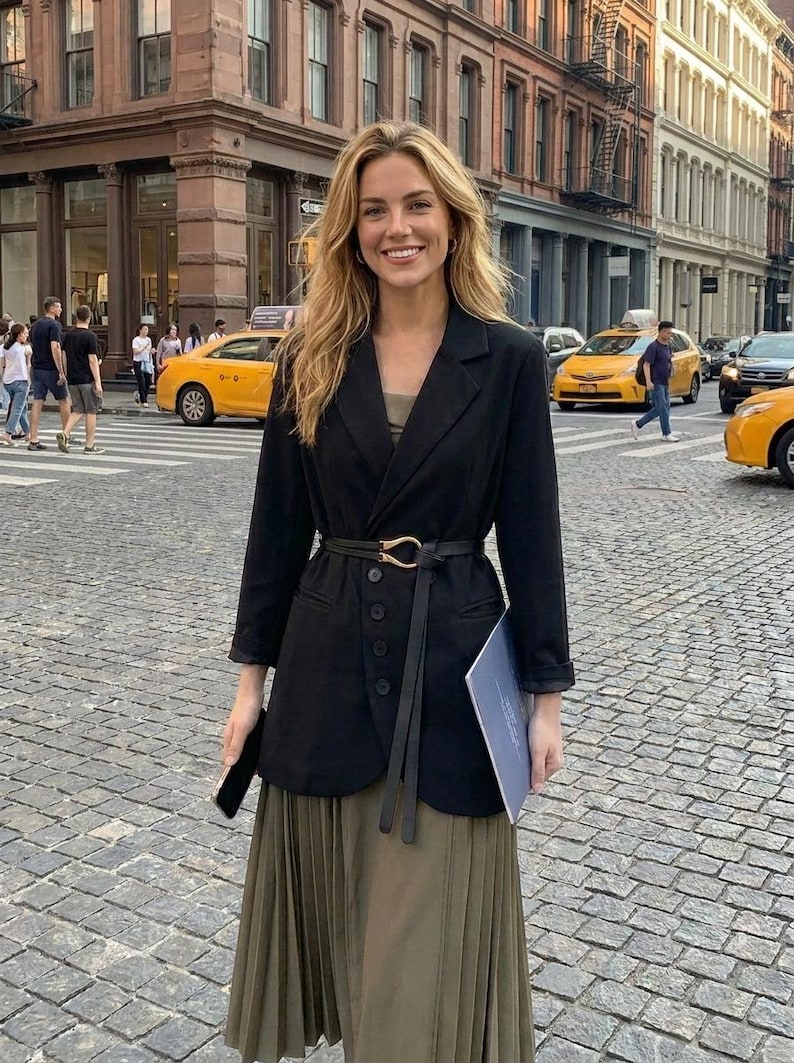 Puede incluir: Una persona con una blazer negra con cintur&oacute;n a juego y una falda plisada larga verde oliva. La blazer tiene cierre de botones y se combina con un top oscuro. El fondo muestra una calle de la ciudad con taxis amarillos y peatones.