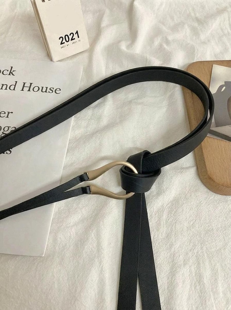 Puede incluir: Cintur&oacute;n negro con hebilla dorada. El cintur&oacute;n est&aacute; hecho de un material liso y presenta un dise&ntilde;o &uacute;nico. El cintur&oacute;n est&aacute; atado con dos extremos largos y colgantes. La hebilla tiene forma curva.
