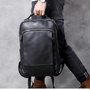 Puede incluir: Una mochila de cuero negro con un diseño rectangular. La mochila tiene un bolsillo frontal con cremallera y un asa superior. La persona lleva vaqueros negros y botas marrones.
