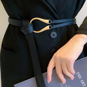 Puede incluir: Cintur&oacute;n negro con hebilla dorada. El cintur&oacute;n est&aacute; envuelto alrededor de una blazer negra. La hebilla tiene un dise&ntilde;o &uacute;nico. El texto del libro dice "THE ART OF".