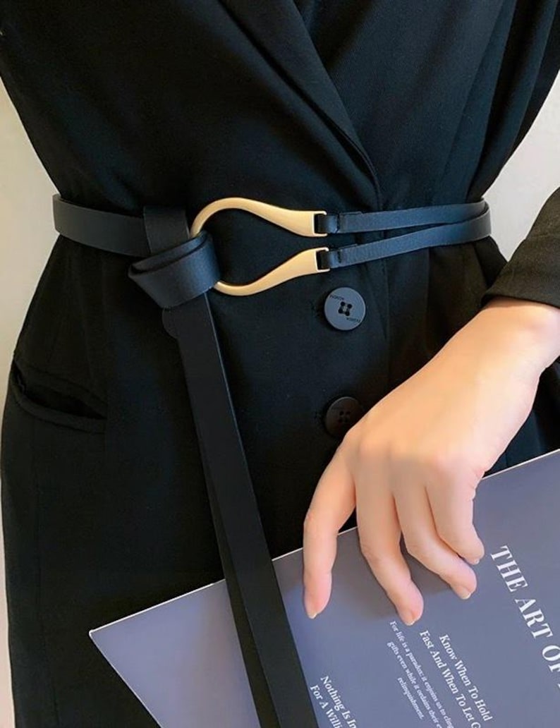 Puede incluir: Cintur&oacute;n negro con hebilla dorada. El cintur&oacute;n est&aacute; envuelto alrededor de una blazer negra. La hebilla tiene un dise&ntilde;o &uacute;nico. El texto del libro dice "THE ART OF".