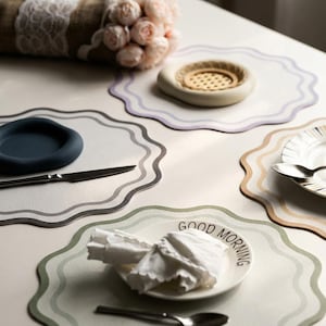 Puede incluir: Una mesa decorada con manteles individuales decorativos en varios colores, incluyendo gris, verde y beige. Se ve un plato con la inscripción "Good Morning", junto con cubiertos y una servilleta. También hay un plato azul pequeño y un cuchillo.