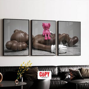 May include: Triptych wall art featuring three framed prints. Each print showcases a brown figure in a relaxed pose, with a pink figure atop one. The figures have white gloves and a white 'X' on the head. The artwork is displayed above a black leather sofa.