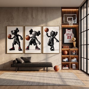 Op de afbeelding: Drie ingelijste wandkunstwerken met zwarte cartoonachtige figuren die basketballen met oranje ballen, boven een zwarte bank. Een houten wandmeubel bevat een wit Bulls 23-shirt, een basketbalring en diverse sportmemorabilia.