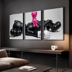 May include: A triptych wall art set featuring a large black character with white gloved hands and X-shaped eyes lying down, with a smaller bright pink character sitting on its chest. The artwork is displayed in three separate black frames.