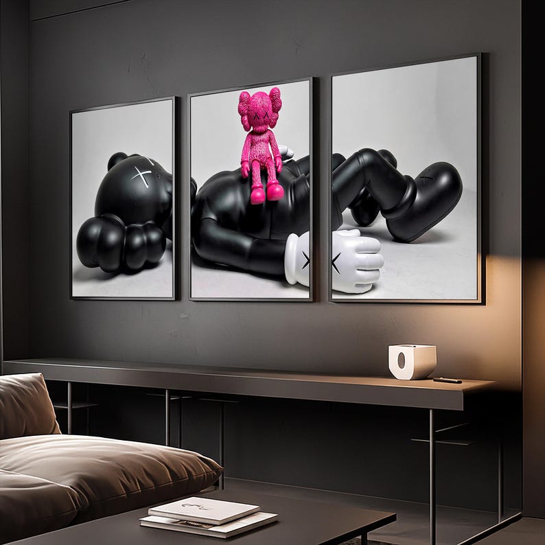 May include: A triptych wall art set featuring a large black character with white gloved hands and X-shaped eyes lying down, with a smaller bright pink character sitting on its chest. The artwork is displayed in three separate black frames.