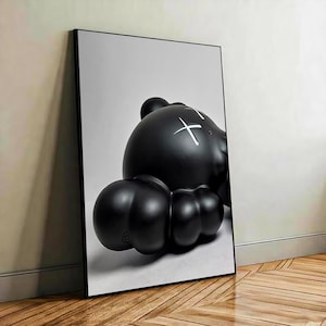 May include: A framed vertical wall art print featuring a glossy black rounded character with a smooth, bulbous form. The character has white cross-shaped markings over its eyes and is set against a plain, light gray background within a thin black frame.