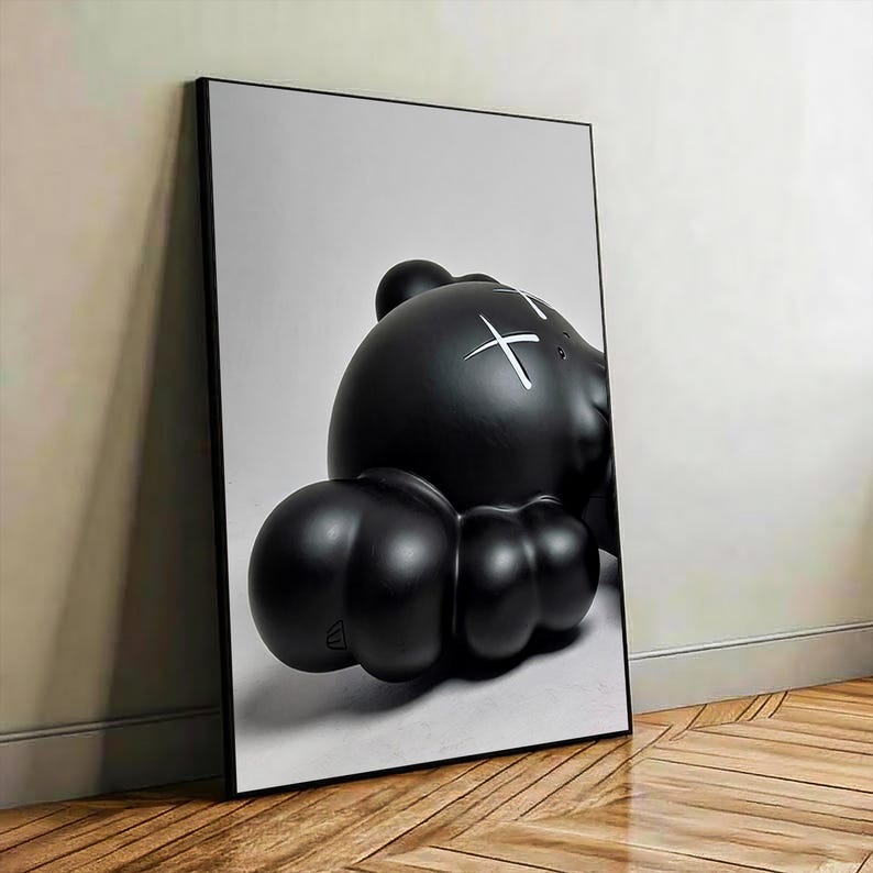 May include: A framed vertical wall art print featuring a glossy black rounded character with a smooth, bulbous form. The character has white cross-shaped markings over its eyes and is set against a plain, light gray background within a thin black frame.