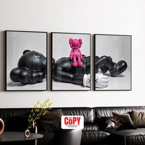 May include: A triptych wall art set featuring a large black KAWS-style character lying down, with a smaller bright pink KAWS-style figure sitting on its chest. The black figure has white gloved hands and X-shaped eyes, while the pink figure is solid.