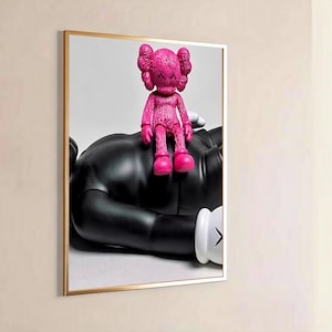 May include: A framed wall art print featuring a bright pink textured KAWS figure sitting on top of a larger, smooth black KAWS figure. The black figure is lying down and has white gloved hands with black X markings on the palms visible in the frame.
