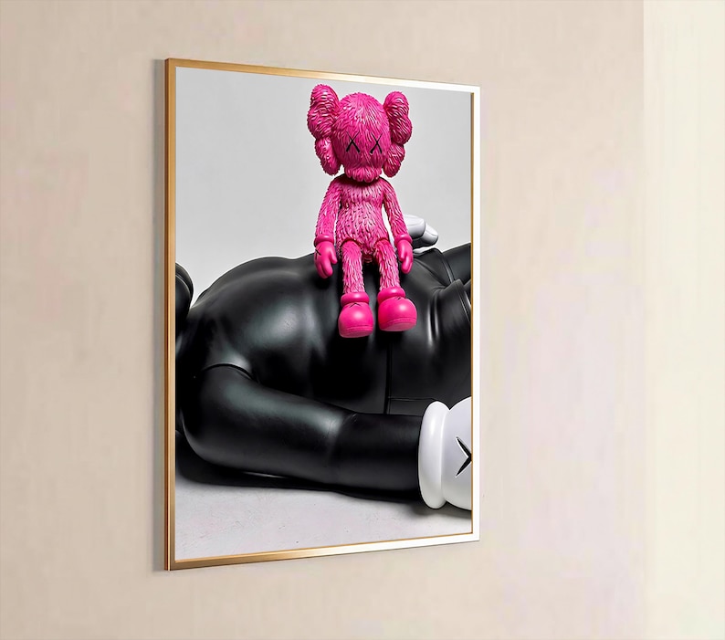 May include: A framed wall art print featuring a bright pink textured KAWS figure sitting on top of a larger, smooth black KAWS figure. The black figure is lying down and has white gloved hands with black X markings on the palms visible in the frame.