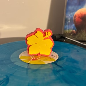 Könnte beinhalten: Ein hellgelber und rosafarbener, hibiskusblütenförmiger Schallplatten-Stabilisator. Der Stabilisator liegt auf einer blau-weiß marmorierten Schallplatte mit einem Mittellabel, das eine gelbe Hibiskusgrafik und den Text Midnight Sun zeigt.