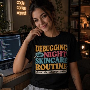 May include: A black t-shirt featuring the colorful text "DEBUGGING IS MY NIGHT SKINCARE ROUTINE" in yellow, blue, and pink, with the smaller text "cleanse cache • patch bugs • glow later" printed in a white rectangular border below the main graphic.
