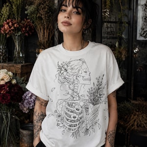 Può includere: Una t-shirt bianca a maniche corte con un disegno a linee nere raffigurante il busto e la testa di uno scheletro adornati con fiori. Lo scheletro è ritratto di profilo mentre tiene un bouquet di fiori di campo, con elementi floreali integrati nella cassa toracica.