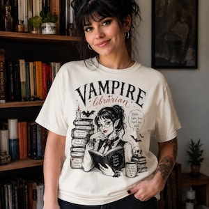 May include: An off-white short-sleeve t-shirt featuring a graphic illustration of a vampire librarian reading a book. The design includes stacks of books, a candle, a skull, a mug, bats, and the text "VAMPIRE librarian" and "Shhh... Late fees hurt my soul."