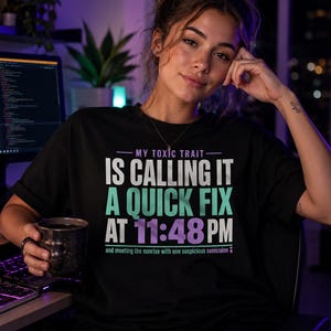 Op de afbeelding: Een zwart T-shirt met korte mouwen met de tekst: MY TOXIC TRAIT IS CALLING IT A QUICK FIX AT 11:48 PM and meeting the sunrise with one suspicious semicolon. De tekst is gedrukt in witte, lichtgroene en paarse letters.