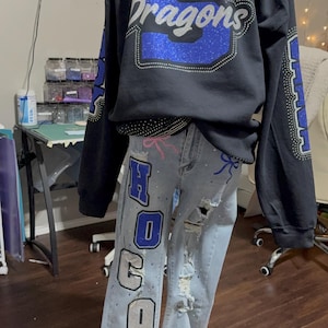Puede incluir: Una sudadera negra y unos vaqueros desgastados sobre un maniquí. La sudadera tiene un diseño de "Dragons" en azul y plata con purpurina. Los vaqueros presentan "HOCO" y un diseño de fútbol americano, con detalles desgastados y acentos rosas.