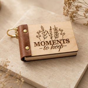 May include: A wooden keychain book with the words "MOMENTS -to keep-" engraved on the front. The book has a brown leather spine with two gold rivets and a gold keychain ring. The front of the book features a floral design.