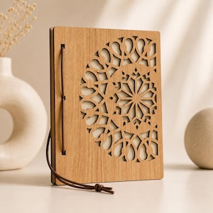May include: A wooden journal with a laser-cut geometric design on the cover. The cover is light brown with a detailed pattern. A leather cord wraps around the journal to keep it closed. The journal is standing upright on a white surface.