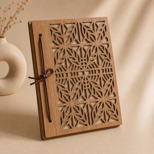 May include: A wooden journal with a laser-cut floral design on the cover. The journal is bound with a brown leather cord. The pages are visible through the cut-out design. The journal is made of light brown wood.