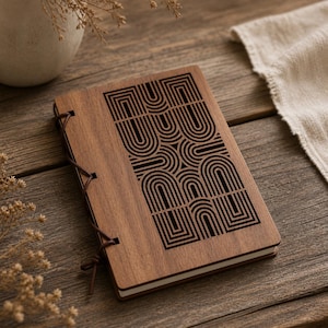 Op de afbeelding: Een houten notitieboek met een geometrisch ontwerp op de kaft. De kaft is gemaakt van donker hout en heeft een zwart, herhalend patroon van gebogen lijnen en rechthoeken. Het notitieboek is gebonden met een bruin leren koord.