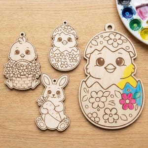 Może przedstawiać: Cztery niedokończone drewniane ozdoby wielkanocne: pisklę z koszem kwiatów, pisklę wykluwające się z jajka, królik trzymający marchewkę i większe pisklę wykluwające się z jajka. Największa ozdoba jest częściowo pomalowana.