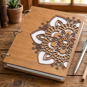 Może przedstawiać: Drewniany notatnik z wycinanym laserowo kwiatowym wzorem na okładce. Notatnik ma jasnobrązowe drewniane wykończenie i srebrne metalowe pierścienie. Kwiatowy wzór jest skomplikowany i szczegółowy, z białym tłem widocznym przez wycięty wzór.