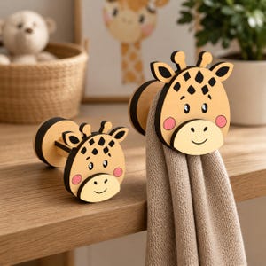 May include: Two wooden giraffe-shaped hooks with black and tan details. The hooks have a smiling face and pink cheeks. One hook holds a tan towel. The hooks are on a wooden surface, with a giraffe picture in the background.