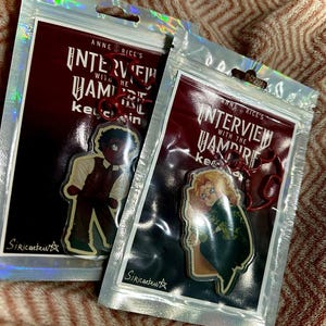 Pode incluir: Dois porta-chaves de acrílico em sacos de plástico holográficos, cada um com uma personagem de Interview with the Vampire de Anne Rice. Um porta-chaves mostra uma figura de cabelo escuro com colete, o outro uma figura loira com casaco verde, ambos com fechos vermelhos.