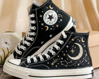 May include: Black high-top sneakers with white laces and soles. The sneakers feature embroidered celestial designs, including moons, stars, and constellations. The Converse All Star logo is visible on the side. A stylish and unique footwear option.