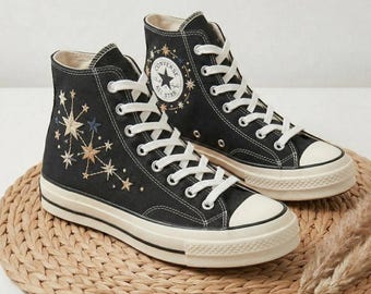 May include: Pair of black high-top sneakers with white laces and a cream-colored sole. The sneakers feature embroidered starburst designs in gold and blue. The Converse All Star logo is visible on the side.