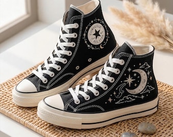 May include: Pair of black high-top sneakers with white laces and soles. One shoe features a star and moon design, the other has the Converse All Star logo. The shoes are on a woven surface.