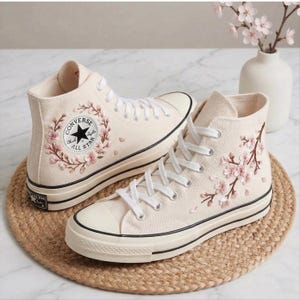 Puede incluir: Zapatillas altas color crema con bordados de flores de cerezo rosas. Las zapatillas tienen cordones blancos, una franja negra en la suela y el logotipo de Converse All Star. Se muestran sobre una estera tejida.