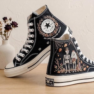 May include: Black high-top sneakers with white laces and soles. One shoe features a "Chuck Taylor All Star" logo with floral embroidery. The other shoe has a skeleton couple design with the text "Skeleton Wedding".