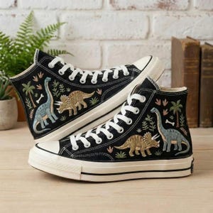Puede incluir: Zapatillas altas negras con cordones y suelas blancas. Las zapatillas presentan bordados de dinosaurios, palmeras y follaje en tonos azules, marrones y verdes. Los zapatos se exhiben sobre una superficie de madera.