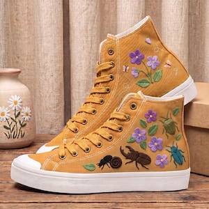 Könnte beinhalten: Ein Paar senfgelbe High-Top-Sneaker aus Canvas mit gestickten violetten Blumen, grünen Blättern und verschiedenen Insekten, darunter Käfer und eine Schnecke. Die Schuhe haben passende gelbe Schnürsenkel, Metallösen und eine weiße Gummisohle.