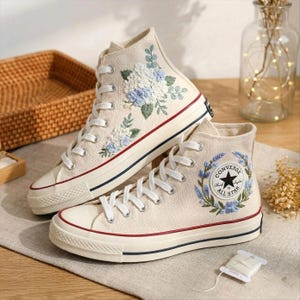 Pode incluir: Par de ténis de lona de cano alto creme com bordado floral. Os ténis apresentam atacadores brancos, uma risca vermelha e azul marinho na sola e o logótipo Converse All Star. São decorados com bordados florais azuis e verdes.