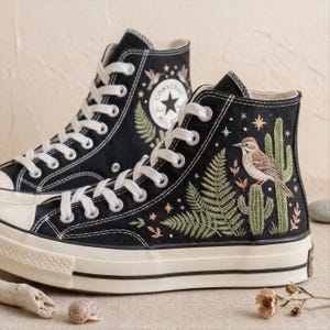 May include: Pair of black high-top sneakers with white laces and soles. The sneakers feature embroidered designs of a bird, cactus, and ferns. The Converse logo is visible on the side.