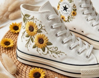 May include: White high-top sneakers with embroidered yellow sunflowers and green leaves. The sneakers have a black stripe along the sole and white laces. The Converse logo is on the side, surrounded by a sunflower wreath.