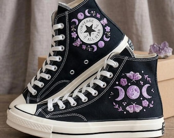 May include: Black high-top Converse Chuck Taylor All Star sneakers with white laces and soles. The sneakers feature embroidered purple floral and moon phase designs. The Converse logo is visible on the side.