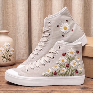 Könnte beinhalten: Ein Paar hellgraue High-Top-Sneaker aus Canvas mit weißer Gänseblümchen-Stickerei und kleinen roten Marienkäfer-Akzenten. Die Schuhe haben weiße Schnürsenkel, weiße Gummikappen an den Zehen und weiße Sohlen.