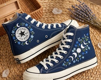 May include: A pair of navy blue high-top sneakers with white laces and soles. The sneakers feature embroidered floral and celestial designs in blue and white. The Converse All Star logo is visible on one shoe.